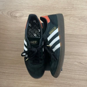 Adidas Spezial sneakers - Snygga Adidas Spezial sneakers i svart med vita ränder och orange detaljer. Skorna har en klassisk design med snörning och en gummisula. Perfekta för en stilren look.