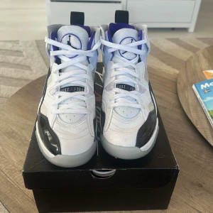 Nike Air Jordans i vitt och svart - Snygga Nike Air Jordans i vitt med svarta och lila detaljer. Skorna har en hög design med snörning och en ikonisk logga på plösen. Perfekta för dig som vill ha en stilren och sportig look.