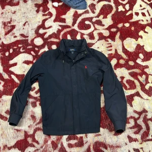 Mörkblå  jacka från Polo Ralph Lauren - Säljer en stilren mörkblå jacka från Polo Ralph Lauren. Jackan har en klassisk design med dragkedja och långa ärmar. Perfekt för en snygg och avslappnad look.