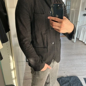 Mörkgrå kavaj från Nudie - Snygg mörkgrå kavaj med knappar framtill och flera fickor. Perfekt för en stilren look. Kavajen har långa ärmar och en klassisk krage.