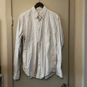 Vit och blå rutig skjorta från GANT - Säljer en klassisk vit och blå rutig skjorta från GANT. Skjortan är tillverkad i 100% bomull och har långa ärmar samt knappar framtill. Perfekt för en stilren look.