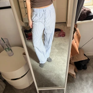 Randiga blå och vita byxor - Säljer ett par bekväma och stilrena byxor i blått och vitt randigt mönster. Byxorna har en loose passform och är perfekta för en avslappnad look. De är tillverkade i ett lätt och luftigt material, vilket gör dem idealiska för varmare dagar.