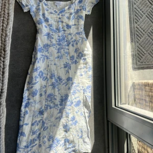 Blommig klänning från & Other Stories - Vit klänning med blått blommönster från & Other Stories. Klänningen har korta ärmar och en smickrande passform med smockdetaljer i midjan. 