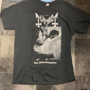 Svart t-shirt med mayhem - Säljer en svart t-shirt med ett stort tryck på framsidan. Trycket har ett mörkt och gotiskt tema med texten 'Pure Fucking Armageddon'. Perfekt för dig som gillar en edgy stil. Passar bra till jeans eller svarta byxor.