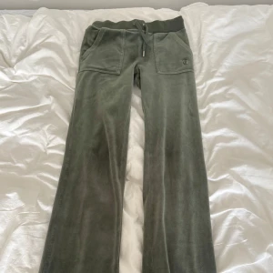 Gröna mjukisbyxor Juicy Couture - Mysiga gröna mjukisbyxor från Juicy Couture i velour med bootcut passform. Byxorna har snörning i midjan, stora fickor fram och bak samt broderad JC-logga på ena fickan. Perfekta för chill dagar eller en avslappnad look. Notera att ena silver snörre fattas en silver knapp. 