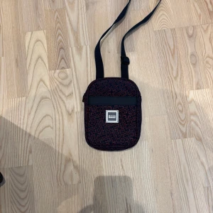 Mörkblå axelväska från Hugo Boss - Snygg mörkblå axelväska från Hugo Boss med röd grafisk logotypmönster över hela väskan. Justerbar axelrem och dragkedja upptill. Perfekt storlek för mobil, plånbok och andra småsaker. Stilsäker detalj med tydlig BOSS-logga framtill.