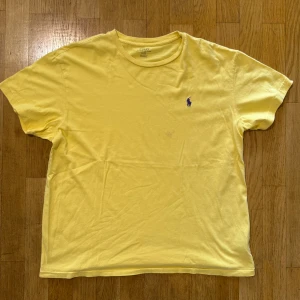 Gul t-shirt från Polo Ralph Lauren - Gul t-shirt från Polo Ralph Lauren med klassisk passform och korta ärmar. T-shirten har en broderad lila logga på bröstet och är gjord i mjuk bomull. Perfekt för dig som gillar stilrena och färgglada plagg.