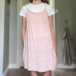 Ljusrosa blommig klänning från Zara - Supersöt ljusrosa kortklänning från Zara med tunna axelband och blommigt mönster i rött och grönt. Klänningen har en luftig passform och är perfekt för varma dagar. Tillverkad i ett mjukt, lätt material som känns skönt mot huden. 