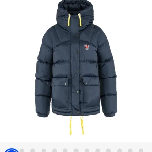 Mörkblå dunjacka från Fjällräven - Snygg mörkblå dunjacka från Fjällräven med huva och gula snören. De är modellen down lite har endast användt den förra vintern och den har inga defekter
