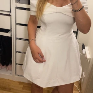 Vit kort klänning med rosett från NA-KD - Supersöt vit kort klänning från NA-KD med offshoulder och stor rosett framtill. Klänningen har en figurnära överdel och utsvängd kjol, perfekt för dig som vill sticka ut. Materialet känns mjukt och har en lätt struktur. Passar perfekt till student eller sommarens alla tillfällen.