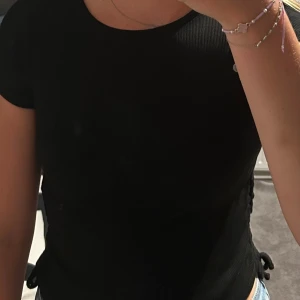 Svart ribbad topp med snörning - En svart ribbad topp med korta ärmar och rund hals. Snygg snörningsdetalj i sidan som ger en extra touch. Barnstorlek men fungerar till xs/s också (väldigt stretchiga material)!💗💕Aldrig använd, då den aldrig kom till användning men prislappen är inte kvar.☺️