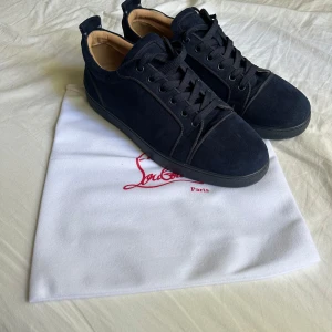 Mörkblå sneakers från Christian Louboutin - Snygga mörkblå sneakers från Christian Louboutin i mocka med klassisk röd sula och svarta snören. Skorna har rund tå och platt sula, samt lyxig beige insida i skinn. Perfekta för dig som vill sticka ut med exklusiv stil och detaljer.