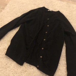 Svart kofta från Next med knappar - En klassisk svart kofta från Next med rund halsringning och snygga knappar längs hela framsidan. Långärmad och i mjukt material som passar perfekt till lager-på-lager-stil. Enkel att matcha med både jeans och kjol.