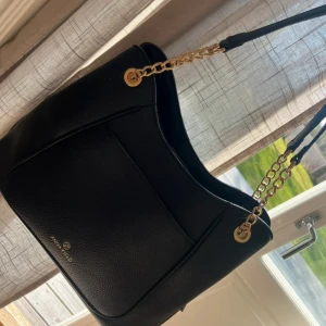 Svart väska - Svart stor väska med guldkedja och märke. Den har 3 stora fack och 2 små fack, ena på utsidan andra på insidan.