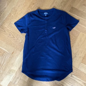 Mörkblå Henley t-shirt från Hollister - Snygg mörkblå Henley t-shirt från Hollister med tre knappar framtill och klassisk rund hals. T-shirten har korta ärmar och är gjord i mjuk bomull. Liten broderad logga på bröstet för en clean look.