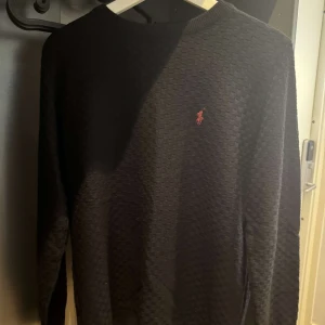 Svart stickad tröja från Ralph Lauren - Säljer en svart stickad tröja från Ralph Lauren med diskret strukturmönster och klassisk röd logga broderad på bröstet. Tröjan har rund halsringning och långa ärmar, perfekt för dig som gillar stilren och tidlös design.
