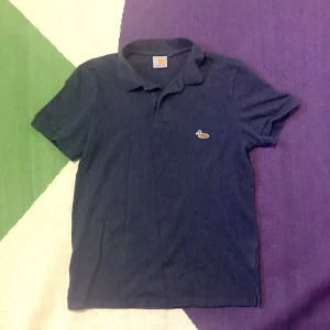 Mörkblå pikétröja från Carhartt - Snygg mörkblå pikétröja från Carhartt med klassisk krage och korta ärmar. Tröjan har en liten broderad anka på bröstet som detalj. Perfekt för dig som gillar enkel och stilren streetwear.