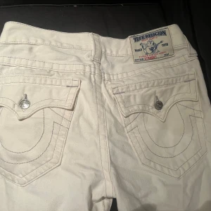 Vita True Religion straight jeans - Säljer ett par helt nya vita jeans från True Religion med klassiska stora bakfickor och snygga silverfärgade knappar. Modellen är straight fit och byxorna har tydliga kontrastsömmar. Perfekta för dig som vill sticka ut med en clean och fräsch look.