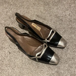 Svarta och silverfärgade pumps El Barato - Unika pumps från El Barato i svart och silver med coola geometriska detaljer. Skorna har fyrkantig tå, låg klack och öppen häl. Tillverkade i skinn med metallicinslag och snygg kontrast mellan färgerna. Perfekta för dig som gillar retrovibbar och statement-detaljer.