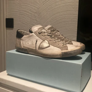 Philippe Model vita sneakers herr 44 - Snygga Philippe Model PRSX Low Man sneakers i vitt och silver med gråa detaljer. Skorna har klassisk snörning med logga på skosnörena, rund tå och platt sula. Tillverkade i skinn och mocka, med svart hälparti och silverdetalj på sidan. Box och dustbag medföljer.