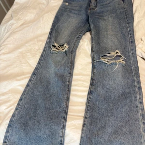 Blå raka jeans med slitna knän - Säljer ett par blå jeans med raka ben och snygga slitningar över båda knäna. Jeansen har klassisk femficksdesign, bälteshällor och är tillverkade i robust denim. Perfekta för en avslappnad och trendig look.