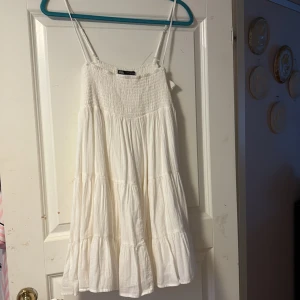 Vit somrig klänning från Zara - Luftig vit klänning från Zara med smock upptill och tunna axelband. Klänningen har volanger och är tillverkad i ett lätt bomullstyg, perfekt för varma dagar. Den har en kort längd och en avslappnad passform.