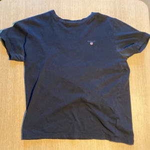 Svart-shirt från Gant - En stilren mörkblå t-shirt från Gant med liten broderad logga och sköld på bröstet. Klassisk rund halsringning och korta ärmar. Tillverkad i mjuk bomull som känns skön mot huden. Perfekt för en clean och enkel look.