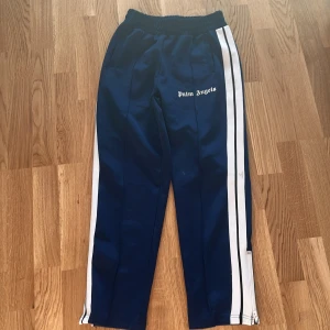 Palm Angels blå träningsbyxor | 170/S - Blå träningsbyxor från Palm Angels med vita sidoränder och logga på låret. Byxorna har elastisk midja med snörning och dragkedja vid benslut. Snygg sportig look och skön passform, perfekta för en streetwear-stil.