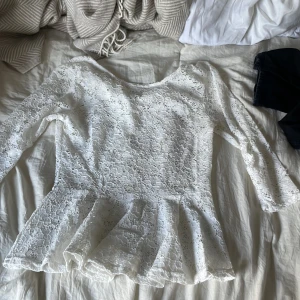 Vit spetsblus från Amisu - Supersöt vit blus från Amisu i spets med blommigt mönster. Modellen har lång ärm, rund hals och volang nertill som ger en peplum-effekt. Perfekt för dig som gillar romantiska och feminina detaljer. Blusen är lätt transparent och har en riktigt fräsch vibe.