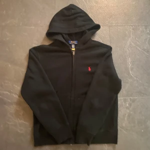 Svart hoodie från Polo Ralph Lauren - Svart hoodie från Polo Ralph Lauren med dragkedja och huva. Klassisk röd broderad logga på bröstet. Tröjan har långa ärmar och är tillverkad i mjukt bomullsmaterial, perfekt för en chill och stilren look.