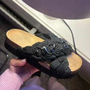 Svarta sandaler med flätad rem och pärlor - Unika svarta sandaler med breda, flätade remmar i stickat material och svarta pärldetaljer. Skorna har en platt korksula med svart botten och öppen tå. Perfekta för dig som vill ha något extra till sommaren. 