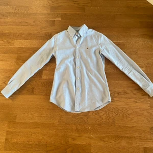 Ljusblå skjorta från Ralph Lauren| XS - Klassisk ljusblå skjorta från Ralph Lauren med broderad logga på bröstet. Tillverkad i mjuk bomull och har en tidlös, normal passform som funkar till många olika stilar. Passar 160-170, Priset kan diskuteras