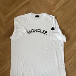 Vit Moncler t-shirt med broderad logga - Snygg vit t-shirt från Moncler med svart broderad logotyp framtill. Klassisk passform med rund hals och korta ärmar. Tillverkad i 100% bomull för en skön och luftig känsla. Perfekt för dig som gillar stilrena och exklusiva plagg.