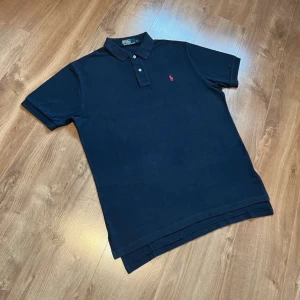 Mörkblå pikétröja från Ralph Lauren - Klassisk mörkblå pikétröja från Ralph Lauren med röd broderad logga på bröstet. Tröjan har korta ärmar, krage och två knappar framtill. Tillverkad i mjuk bomull som känns skön mot huden. Perfekt för en clean och stilren look.
