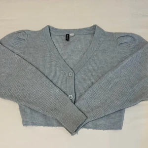 Ljusgblå kofta  - Superfin ljusgrå croppad kofta från Divided med ribbad struktur och v-ringning. Koftan har tre knappar framtill, långa ärmar med lätt puff vid axlarna och är stickad i mjukt material. Perfekt att slänga över en topp för en chill look.