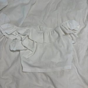 Vit offshoulder croppad topp med volang - Superfin vit croppad topp med offshoulder och bred volang runt axlarna. Ribbad struktur i tyget ger en snygg detalj. Perfekt för dig som gillar en söt och trendig stil.