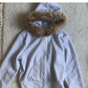 Grå hoodiejacka med fuskpäls från Runway Studio - Grå hoodiejacka från Runway Studio med mjuk fuskpäls runt huvan och glittrig text på ryggen. Jackan har dragkedja framtill, två fickor och är långärmad💋