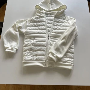 Vit cardigan jacka med huva - Snygg vit cardigan med quiltade partier och dragkedja framtill. Jackan har huva, ribbade muddar och två fickor med dragkedja. Perfekt för dig som gillar en clean stil. Materialet är mjukt och känns skönt mot huden.