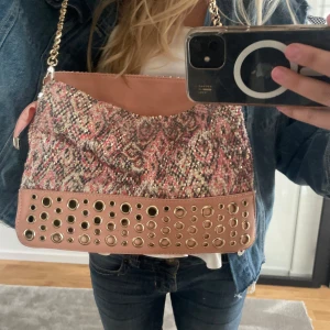 Mönstrad axelväska med kedja och nitar - Axelväska med rosa och beige mönstrad textil, glittriga detaljer och ljusrosa skinnimitation. Väskan har guldfärgad kedjerem, nitar i metall på nederdelen och dragkedja på baksidan. Mått: höjd 22cm, längd: 30 cm , bredd : 10cm