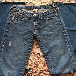 True Religion blå bootcut jeans - Snygga blå jeans från True Religion med klassiska kontrastsömmar och ikoniska broderade bakfickor. Modellen har bootcut-ben och låg midja. Jeansen är i denim med slitningar framtill för en cool, avslappnad look som är med i själva modellen. Köpta för 1tusen i vinted men säljer för 900kr, priset kan diskuteras! Storlek 27 men skulle säga att de går för xs/s