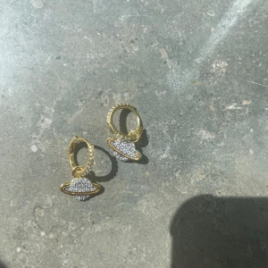 Guldörhängen ZEND DETAILS - Snygga örhängen i guldton med hängande planetmotiv täckt av små glittrande stenar. Runda hoops med extra bling längs hela ringen, perfekta för att ge din look en cool och unik touch. Passar dig som gillar statement-smycken och vill sticka ut.