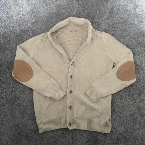 Beige kofta från Massimo Dutti - Snygg beige kofta från Massimo Dutti med stickad struktur, stora knappar och coola mockadetaljer på armbågarna. Koftan har långärmad passform och bred krage som ger en chill och stilren look. Hör av er vid minsta lilla fundering