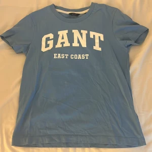 Blå GANT t-shirt med tryck XS - Snygg ljusblå t-shirt från GANT i bomull med vit text 'GANT EAST COAST' på bröstet. Klassisk rund halsringning och korta ärmar. Perfekt för en avslappnad och sportig stil.