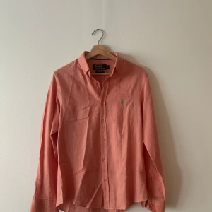 Ralph lauren skjorta - Snygg korallfärgad skjorta från Polo Ralph Lauren i slim fit-modell. Skjortan har klassisk krage, långa ärmar och knäppning framtill. Broderad grön logga på bröstet ger en stilren touch. Perfekt för dig som gillar en fräsch och färgstark 