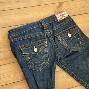 True Religion bootcut jeans blå - Säljer ett par ikoniska blå jeans från True Religion med snygga kontrastsömmar och stora fickor bak med lock och knappar. Modellen är lågmidjad och bootcut, vilket ger en schysst siluett. Klassisk denim med orange detaljer och cool etikett bak.