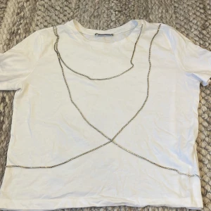 Vit t-shirt med kedjedetalj från Zara - Vit t-shirt från Zara med korta ärmar och rund hals. Framsidan är dekorerad med en glittrig silverkedja. Helt vit baksida. Storlek S. Kedjorna är inte helt fastsydda i T-shirten utan hänger fritt. 