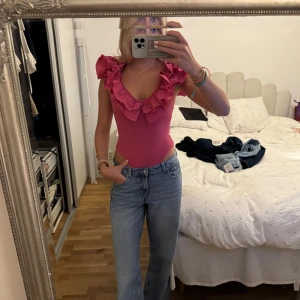 Rosa volangtopp med djup v-ringning - Supersöt rosa topp med djup v-ringning och stora volanger längs axlar och halsringning. Tight passform och ärmlös design, perfekt för dig som vill sticka ut. Materialet är stretchigt och följsamt, vilket gör den både bekväm och snygg.