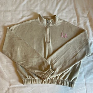 Beige half zip sweatshirt med LA-brodyr - Beige sweatshirt med half zip och broderad LA-text i rosa på bröstet. Tröjan har hög krage, långa ärmar och resår i midjan och vid ärmsluten. Perfekt för en chill och avslappnad stil. Materialet känns mjukt och skönt, troligen bomull eller bomullsblandning.