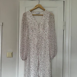 Blommig långklänning med v-ringning - Superfin långklänning från NA-KD i ljus färg med små blommor. Klänningen har v-ringning, knappar framtill och långa, transparenta ärmar. Perfekt för vår och sommar när du vill känna dig extra söt och romantisk.