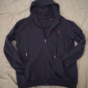 Marinblå hoodie från Polo Ralph Lauren - Riktigt snygg marinblå hoodie från Polo Ralph Lauren med dragkedja och huva. Tröjan har det klassiska röda broderade logotypen på bröstet, snörning i huvan och två fickor framtill. Tillverkad i mjuk bomull för en skön känsla. Om du har frågor så tveka inte eller om du vill ha fler bilder ! Priset är inte hugget i sten !